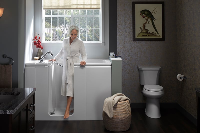 Kohler Walk-In Tub with easy-access door, handheld showerhead, and safety features, shown with senior woman entering for a safe bathing experience