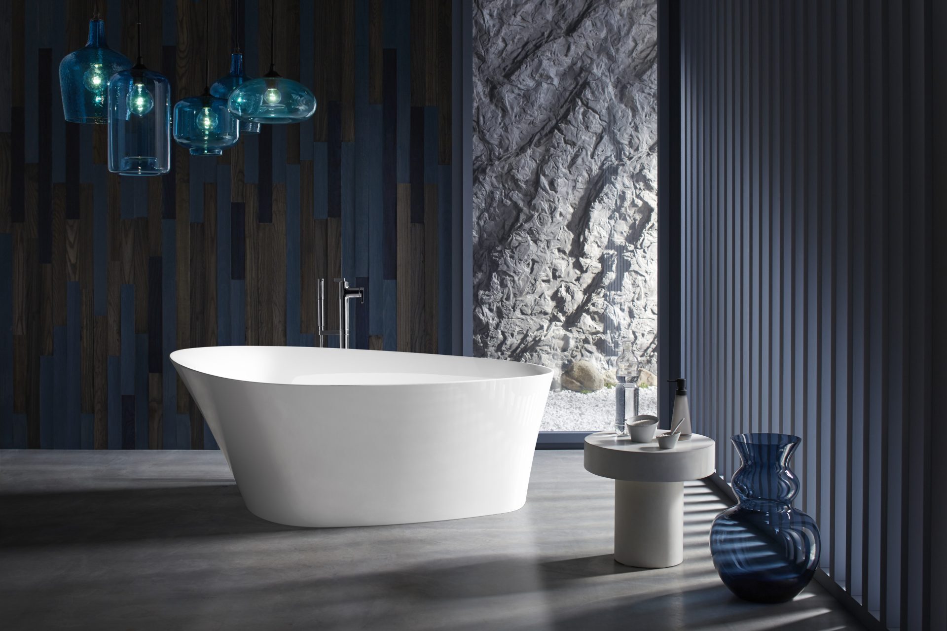 Modern freestanding Kohler soaking tub in a luxury bathroom with dark wood accents, stone wall backdrop, and contemporary lighting.
