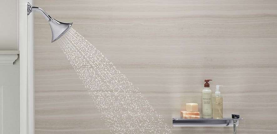 Modern chrome showerhead with running water in a bathroom featuring light stone-look wall panels and a built-in shelf with toiletries.